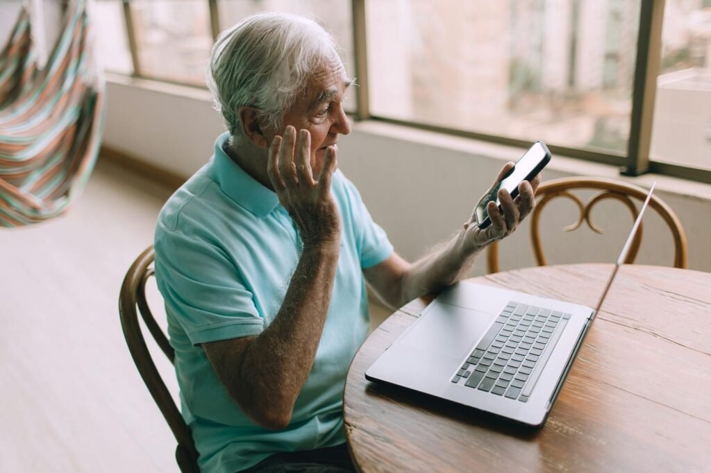 an older man sitting at a table with a laptop and cell phone