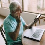 an older man sitting at a table with a laptop and cell phone